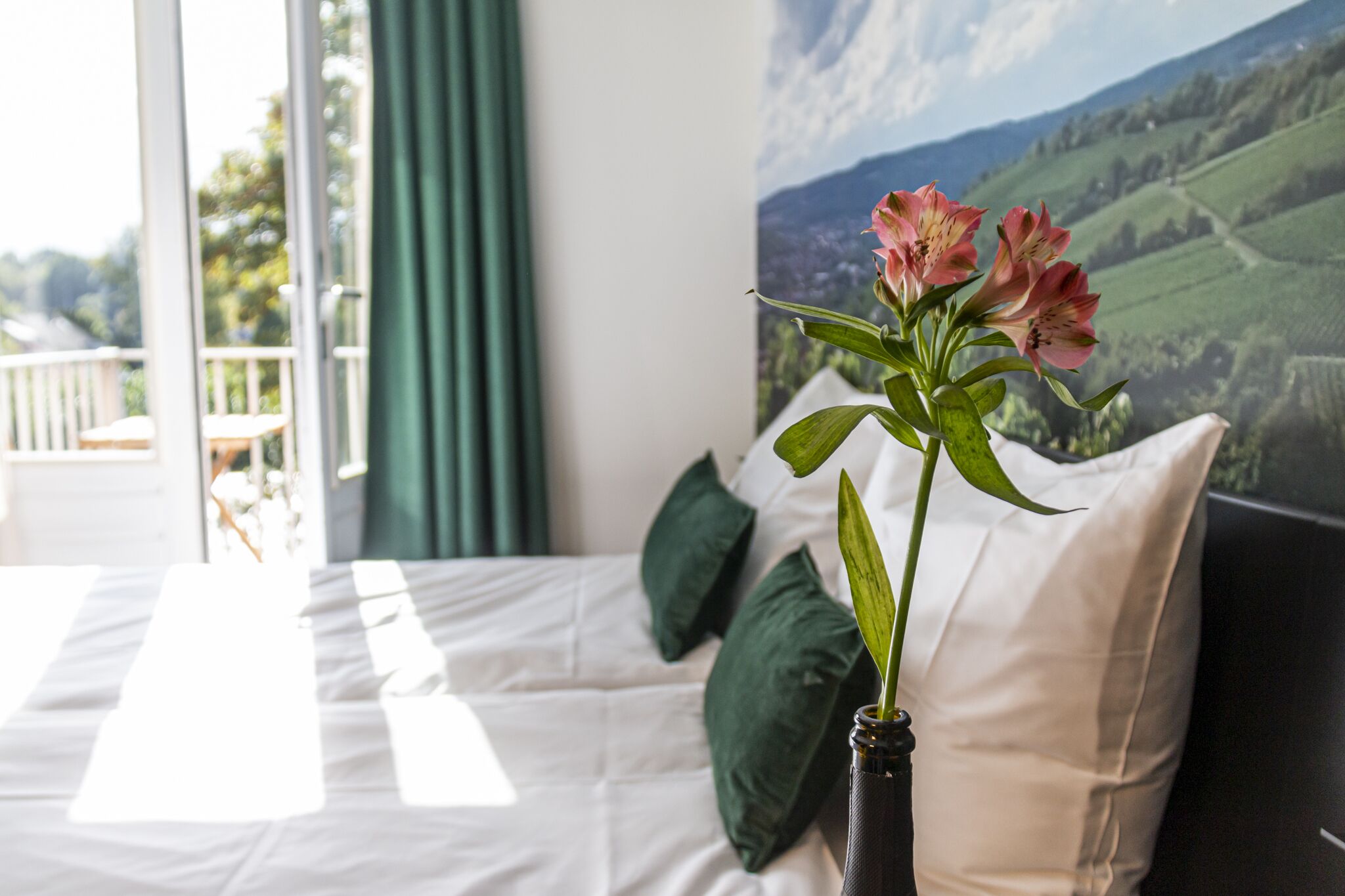Une chambre à coucher avec un lit, des oreillers verts, un vase rempli de fleurs et une grande fenêtre donnant sur un paysage.