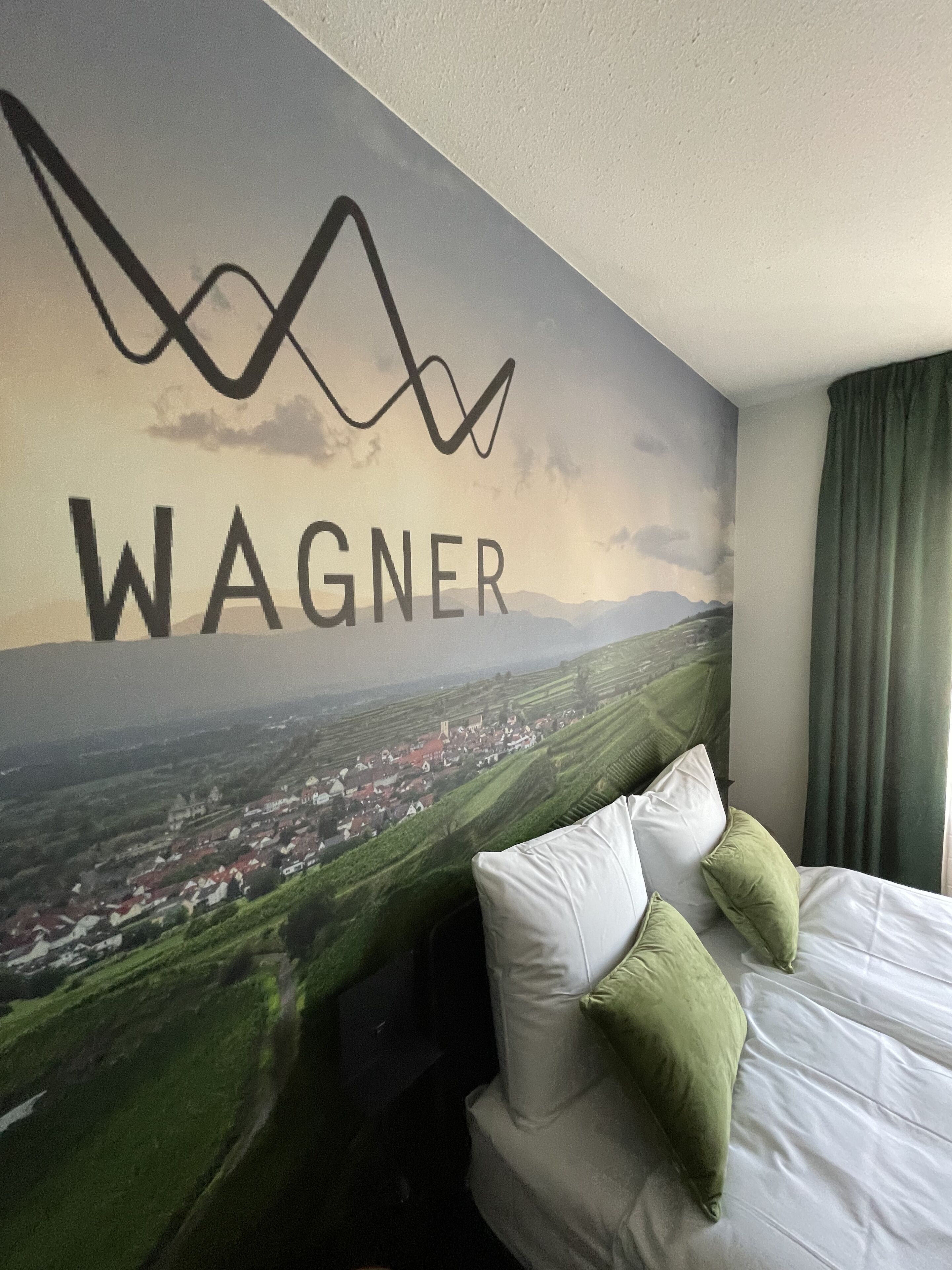 Hotelkamer met een bed, kussens en een muurschildering van een landschap, met de tekst 'WAGNER' op de muur.