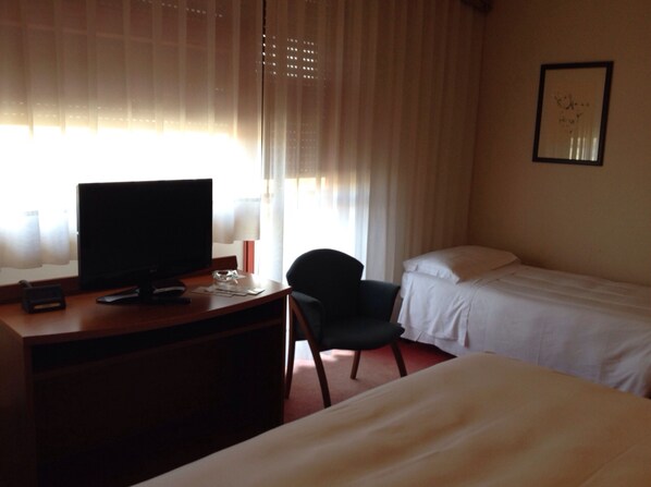 A hotel room with a bed, a desk with a computer monitor, a chair, and a window with curtains.