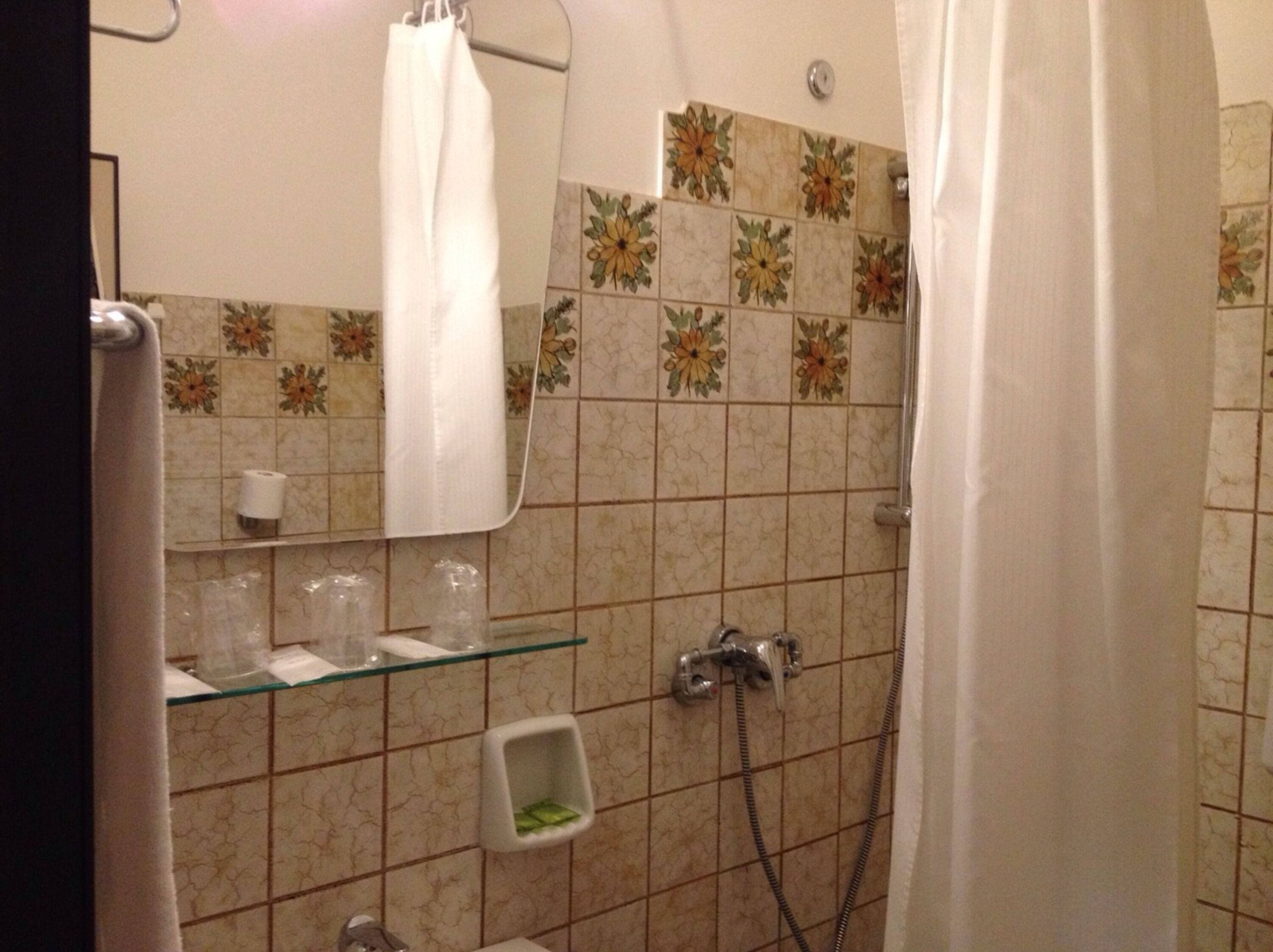 A bathroom with a shower, a glass shelf with toiletries, and a floral tile backsplash.