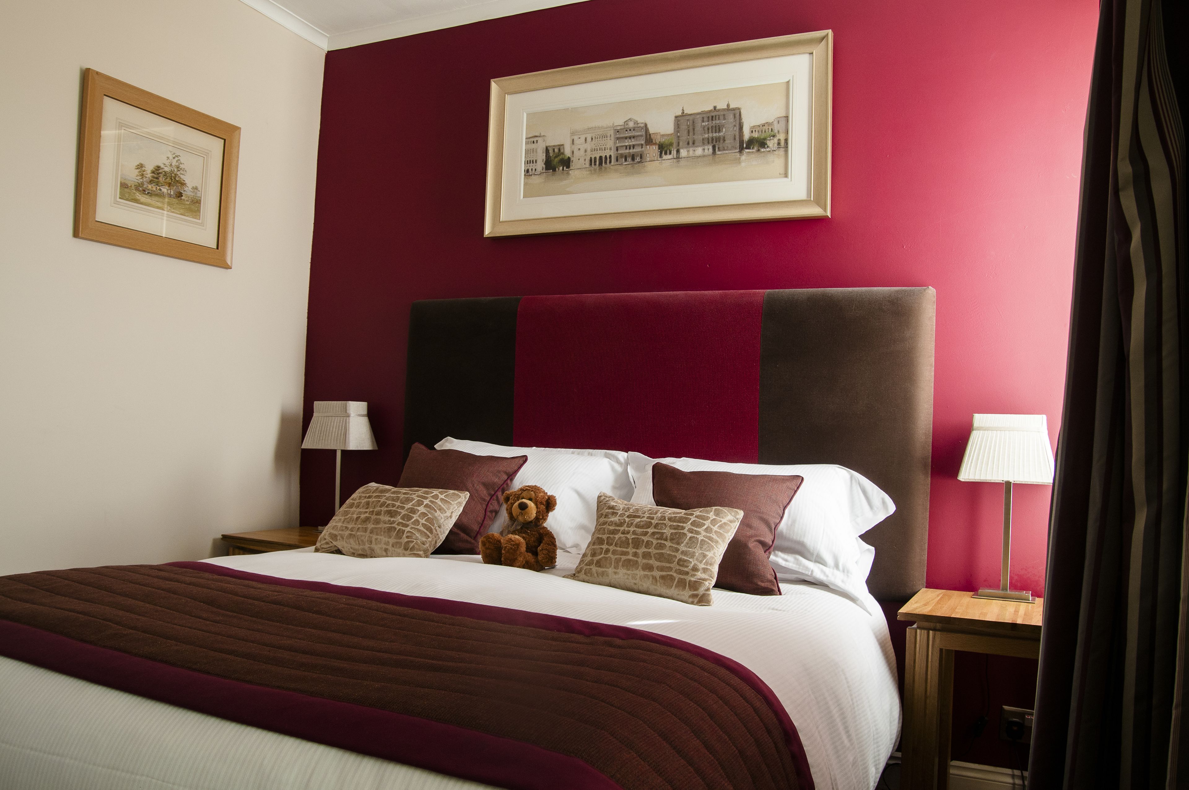 Una habitación de hotel con una cama grande, paredes rojas, dos cuadros enmarcados y una lámpara de noche.