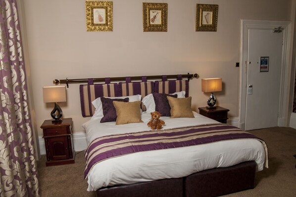 Una cama bien hecha con una colcha a rayas, dos almohadas y un osito de peluche. Hay dos mesitas de noche con lámparas, una puerta y tres cuadros enmarcados en la pared.