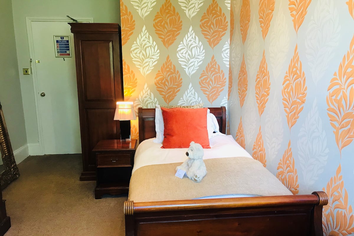 A neatly made bed with a teddy bear, a nightstand with a lamp, and a wooden wardrobe in a hotel room.
