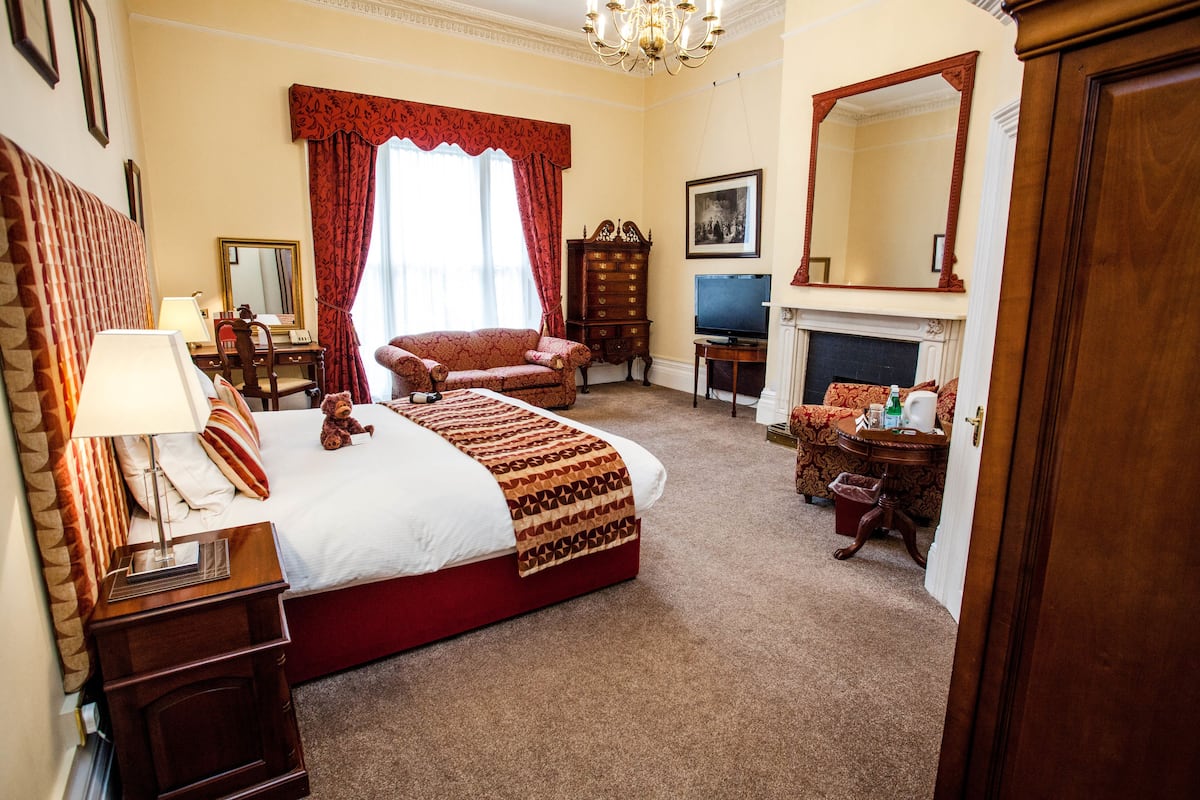 A hotel room with a large bed, a desk, a chair, a television, a fireplace, and a mirror.