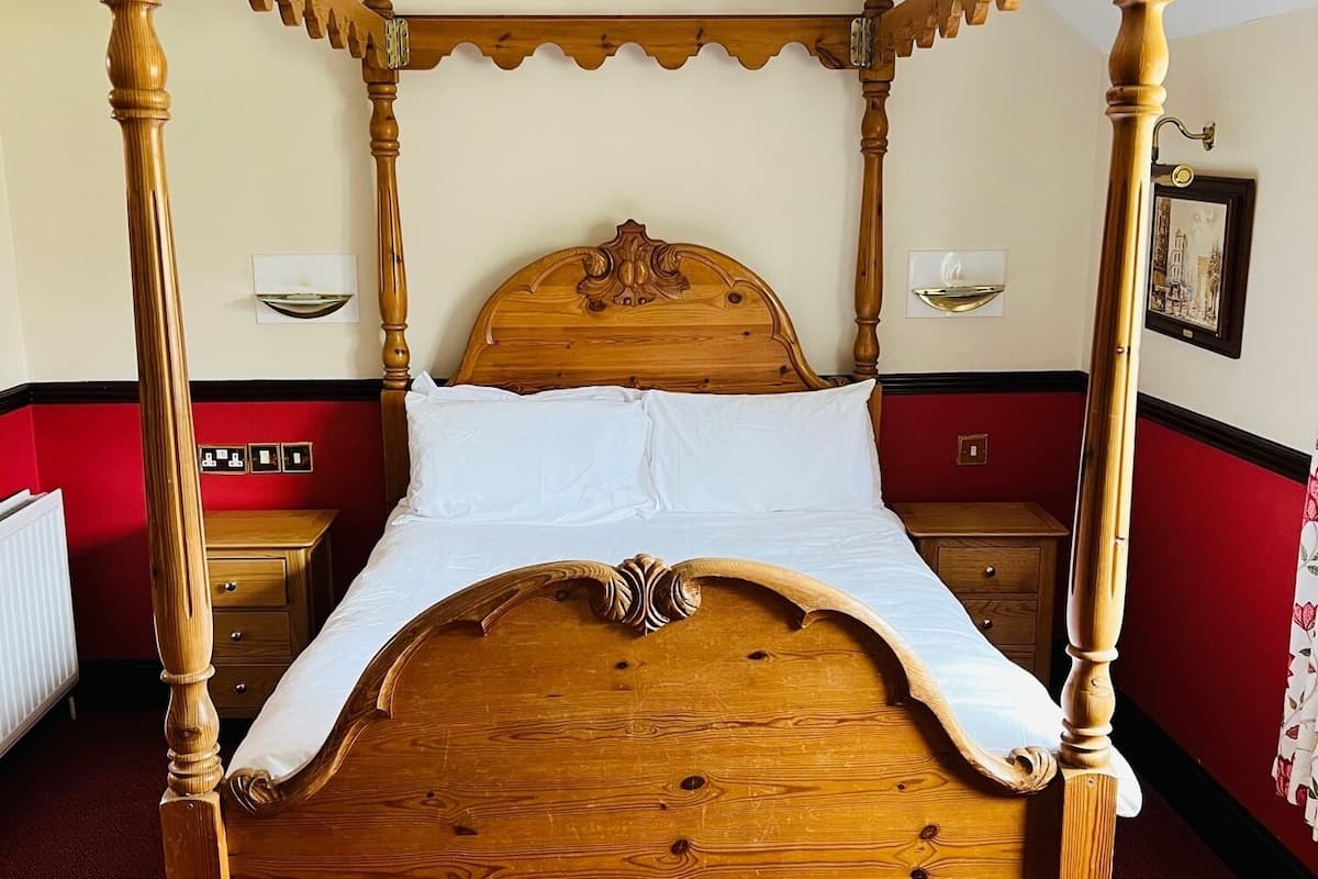 A four-poster bed with a wooden headboard and footboard, a red carpeted floor, and a white wall with a picture frame.