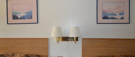 Habitación de hotel con dos camas, una mesita de noche con lámpara, teléfono y reloj. Hay dos cuadros en la pared.