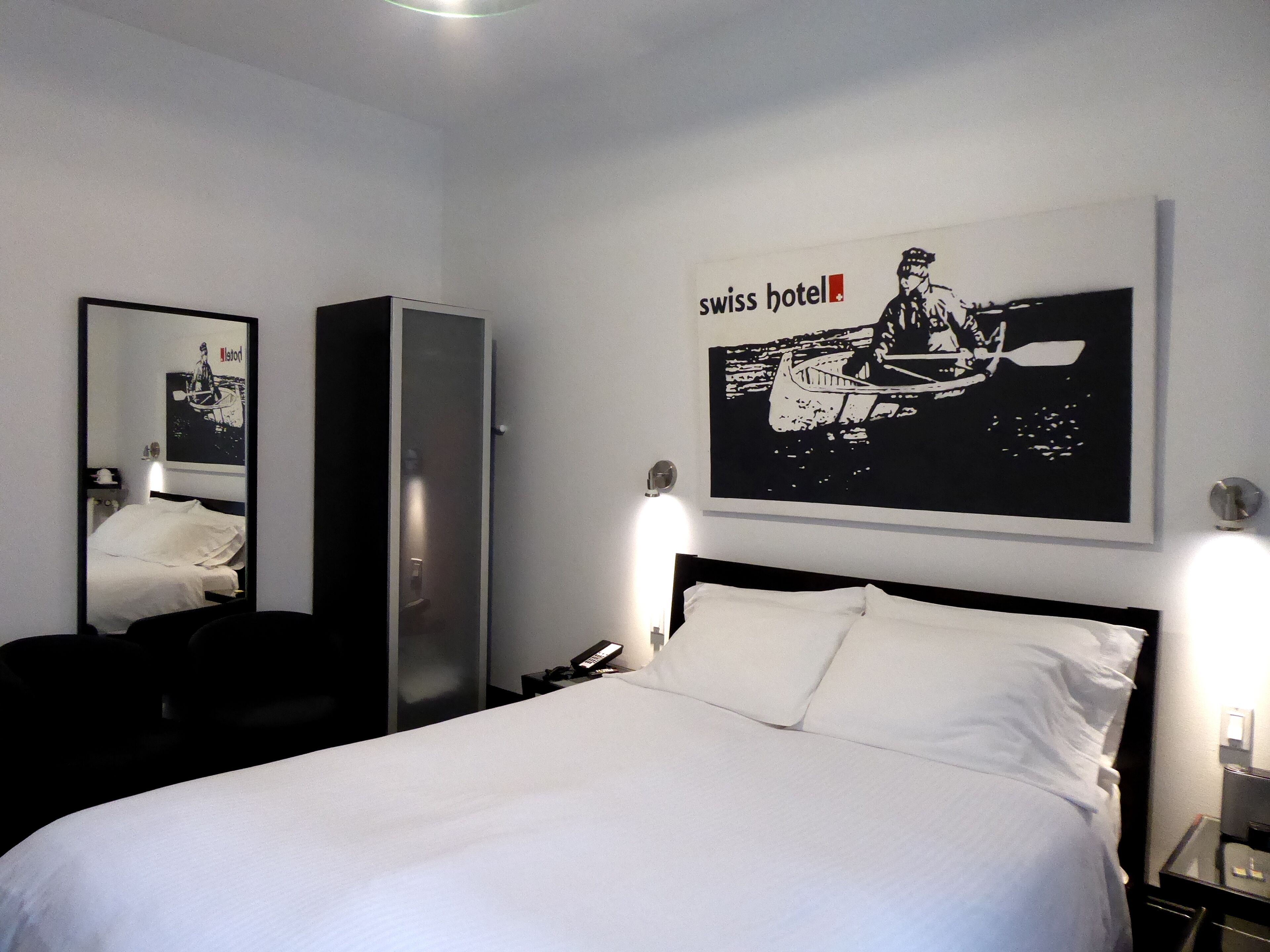 Ein Hotelzimmer mit Bett, Kleiderschrank, Spiegel und einer Wand mit dem Swiss Hotel Logo.