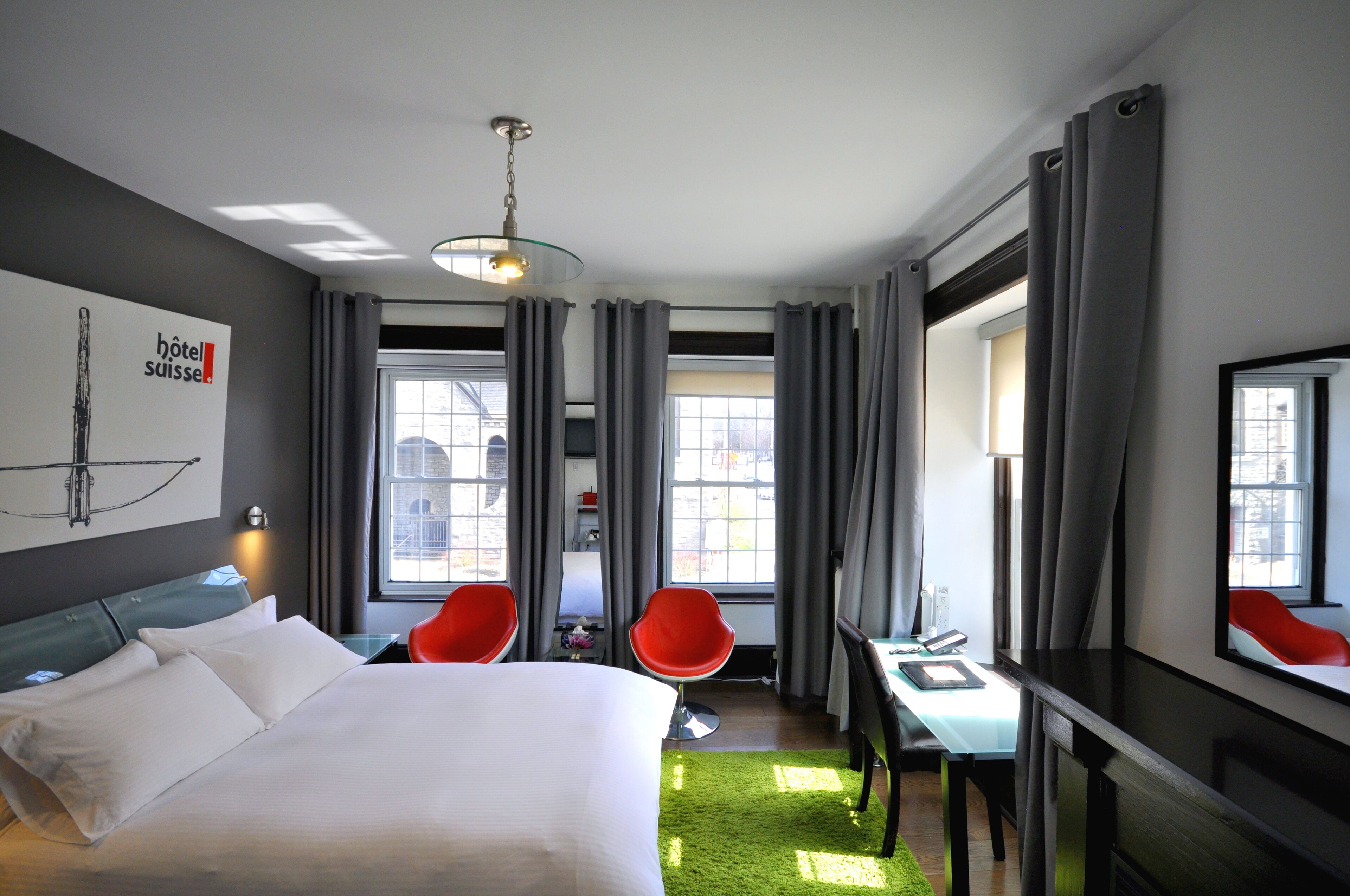 Une chambre d’hôtel avec un lit, deux fauteuils rouges, un bureau et une grande fenêtre.