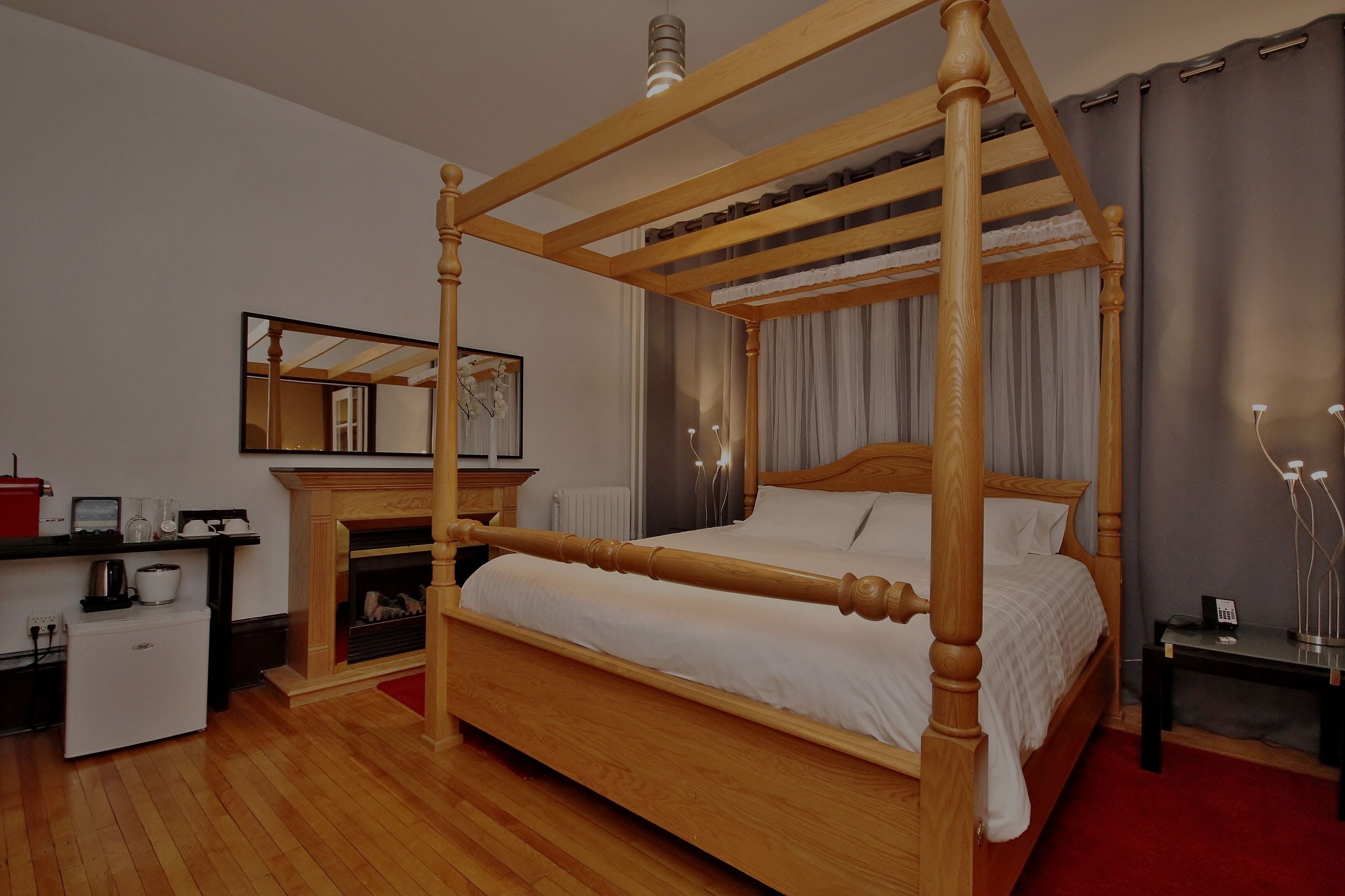 Una cama con dosel de madera, una mesita de noche pequeña, una chimenea, un espejo y una alfombra roja.