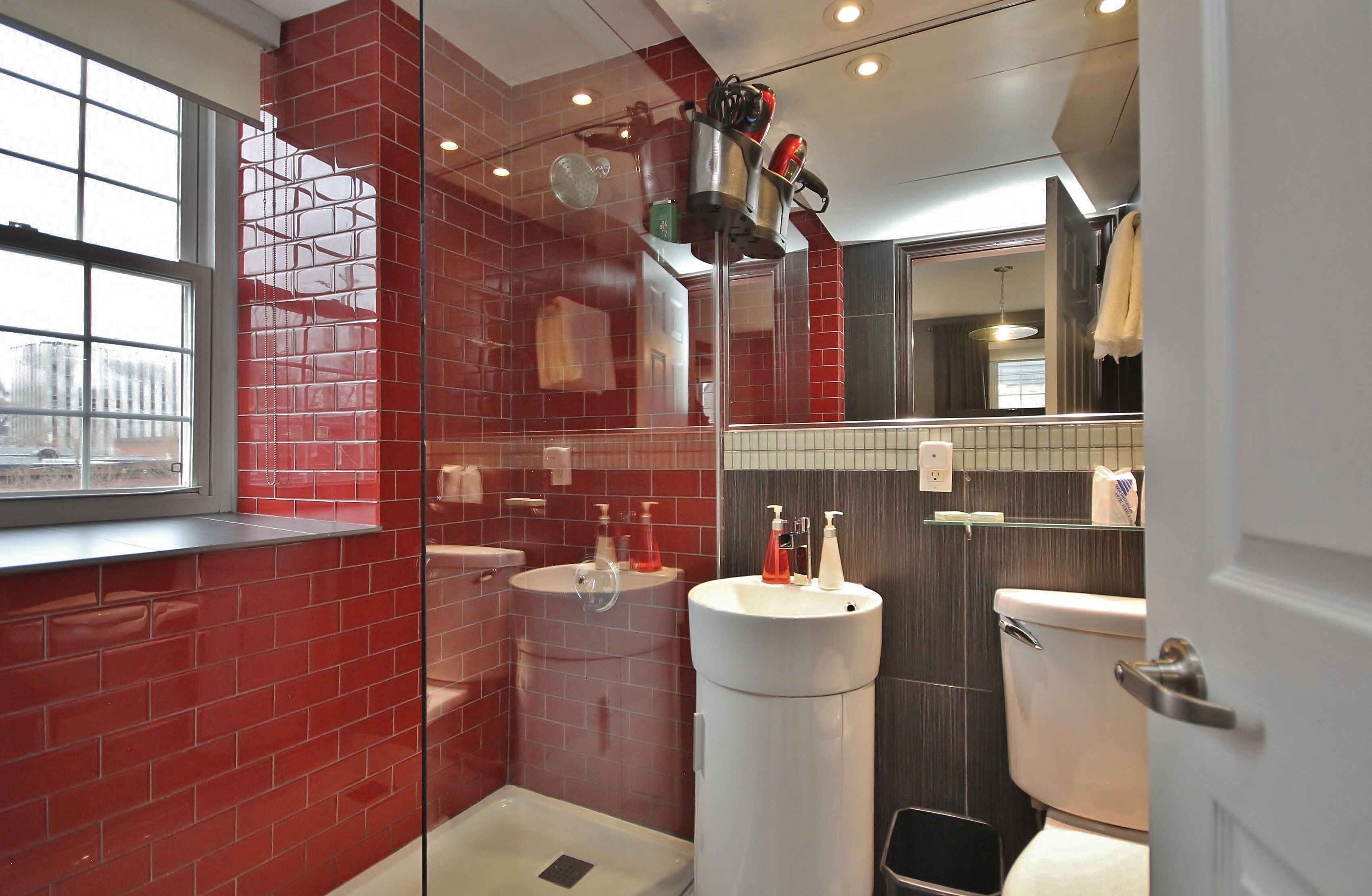 A modern bathroom with red brick walls, a window, two sinks, a toilet, and a large white trash bin.
