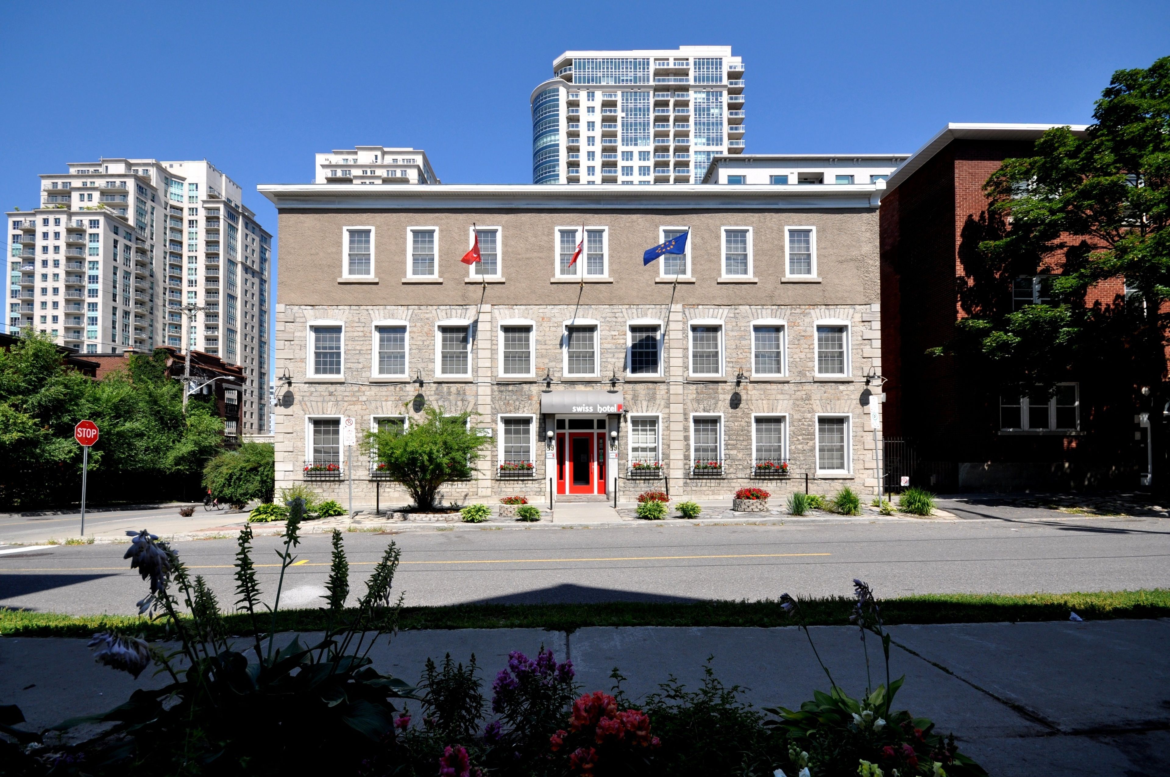Ein zweistöckiges Gebäude mit rotem Eingang, der Beschilderung 'Swiss Hotel' und Flaggen der Vereinigten Staaten, Kanadas und der Europäischen Union.
