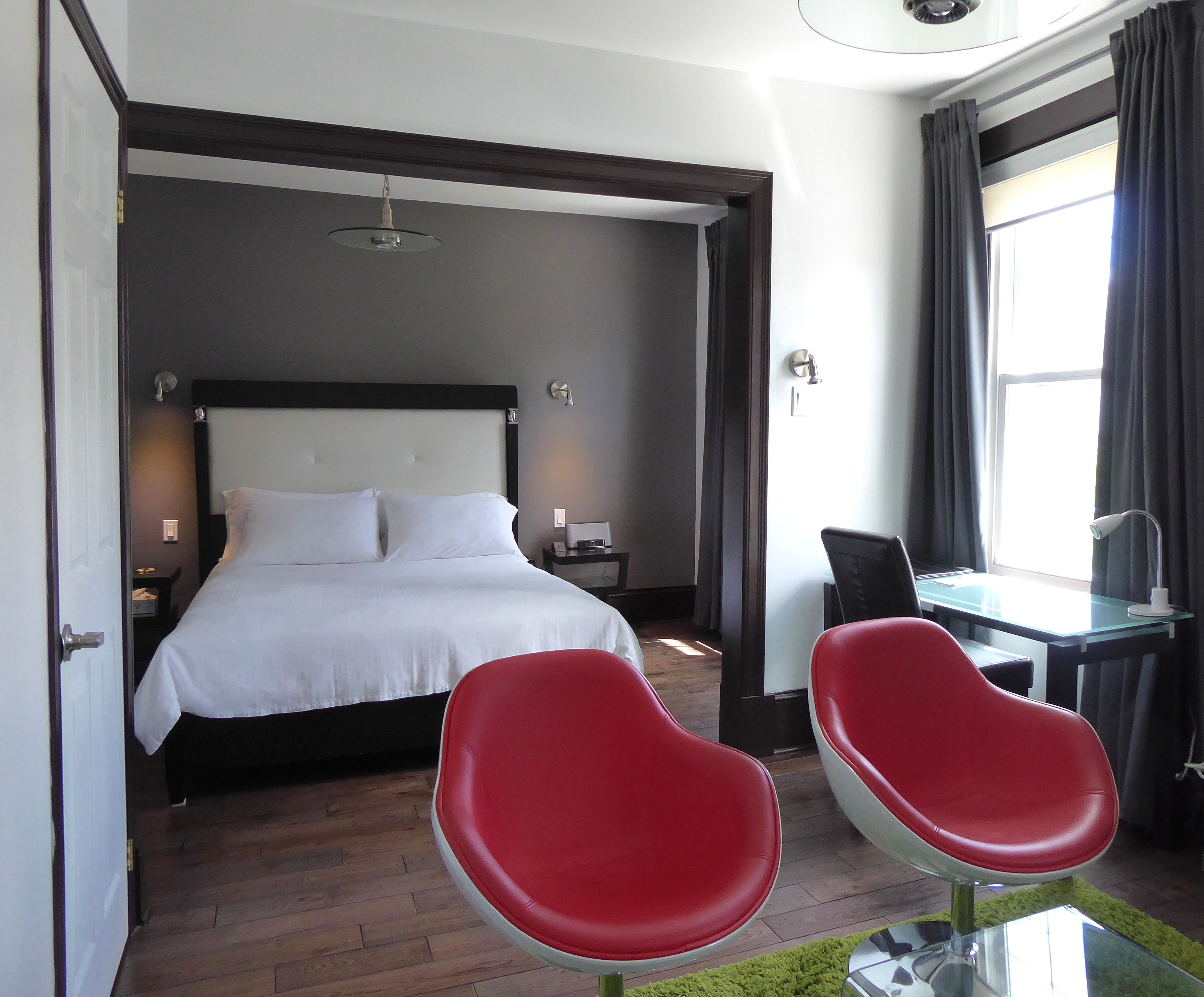 Une chambre d’hôtel avec un lit, un bureau, deux chaises rouges, une fenêtre avec des rideaux et une table de chevet avec une lampe.