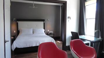 Une chambre d’hôtel avec un lit, un bureau, deux chaises rouges, une fenêtre avec des rideaux et une table de chevet avec une lampe.