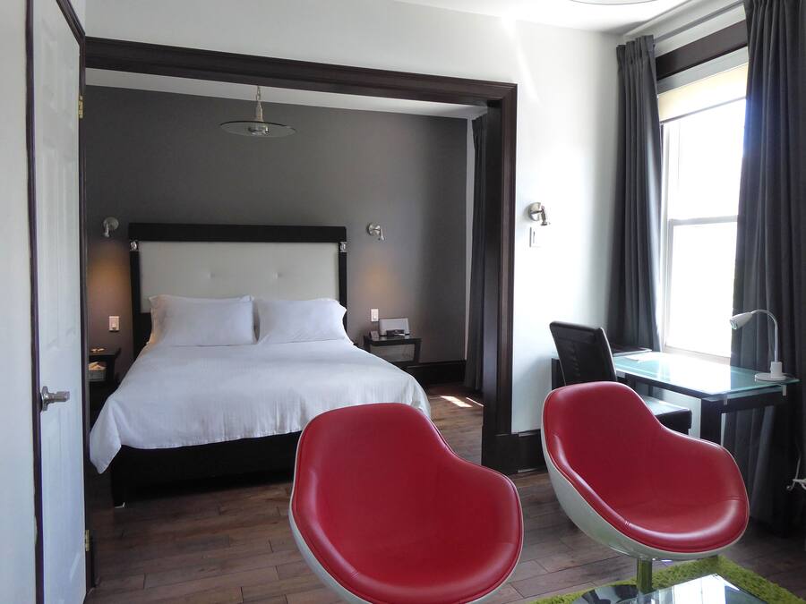 Une chambre d’hôtel avec un lit, un bureau, deux chaises rouges, une fenêtre avec des rideaux et une table de chevet avec une lampe.