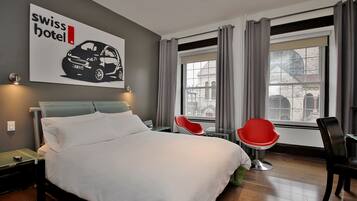 A modern hotel room with a large bed, two red chairs, a desk, and a window with a view of buildings.