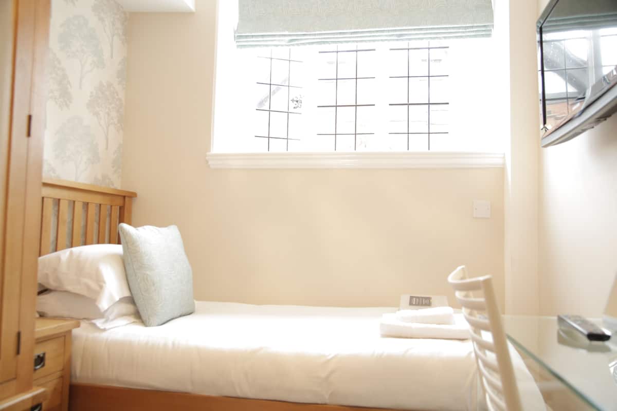 A bedroom with a single bed, a nightstand, a chair, a window with blinds, and a small table with a remote control.