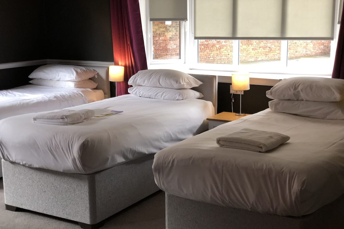 A hotel room with two single beds, each with a pillow and a folded towel. There are bedside lamps and a window with a view of a brick wall.