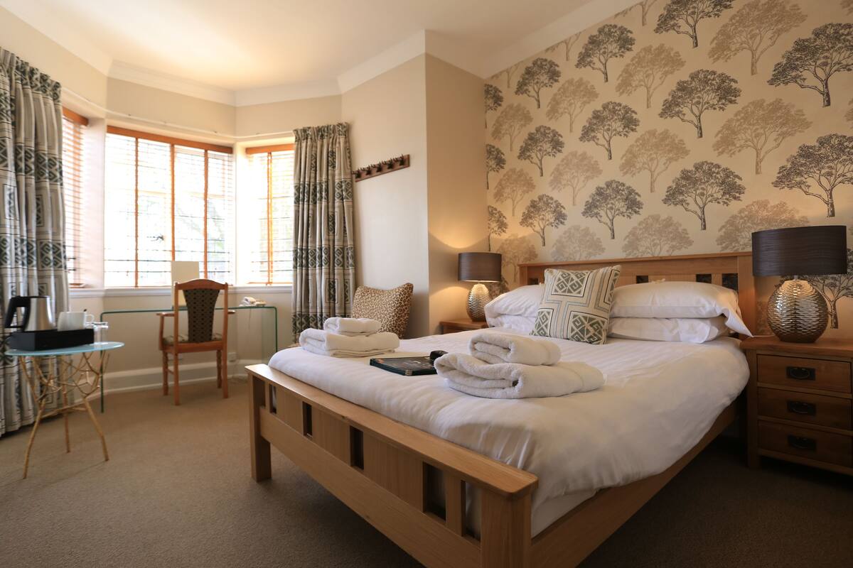 A neatly arranged bedroom with a large bed, a bedside table with lamps, a chair, and a small table with a kettle and cups.
