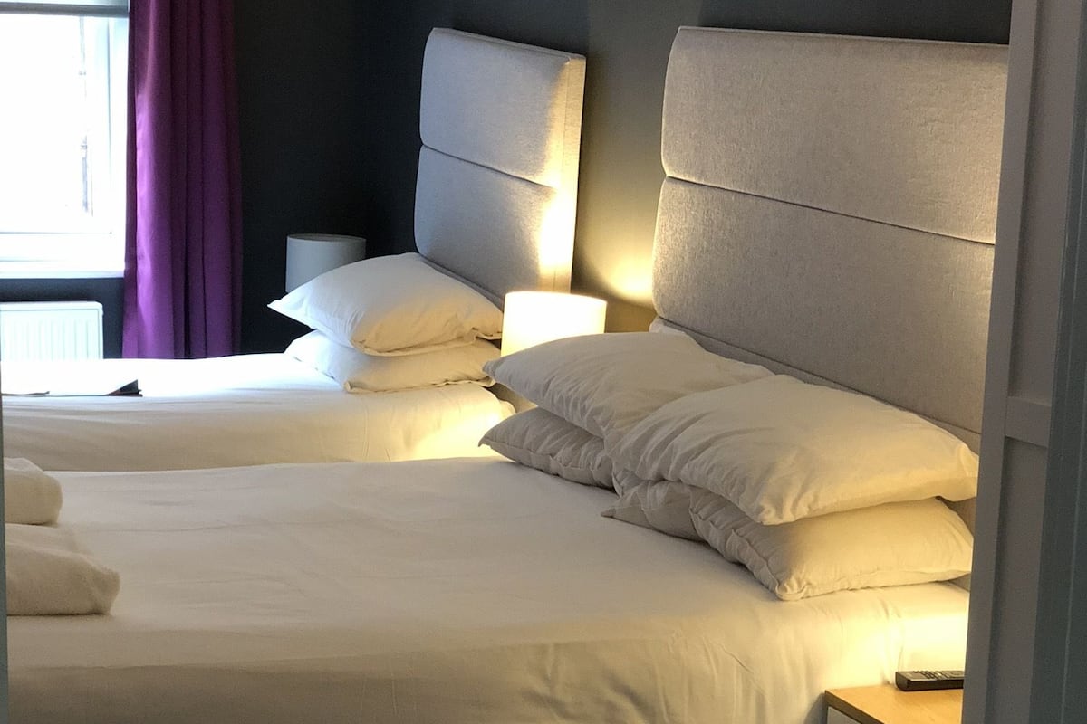 A hotel room with two beds, a nightstand, a lamp, and a window with curtains.