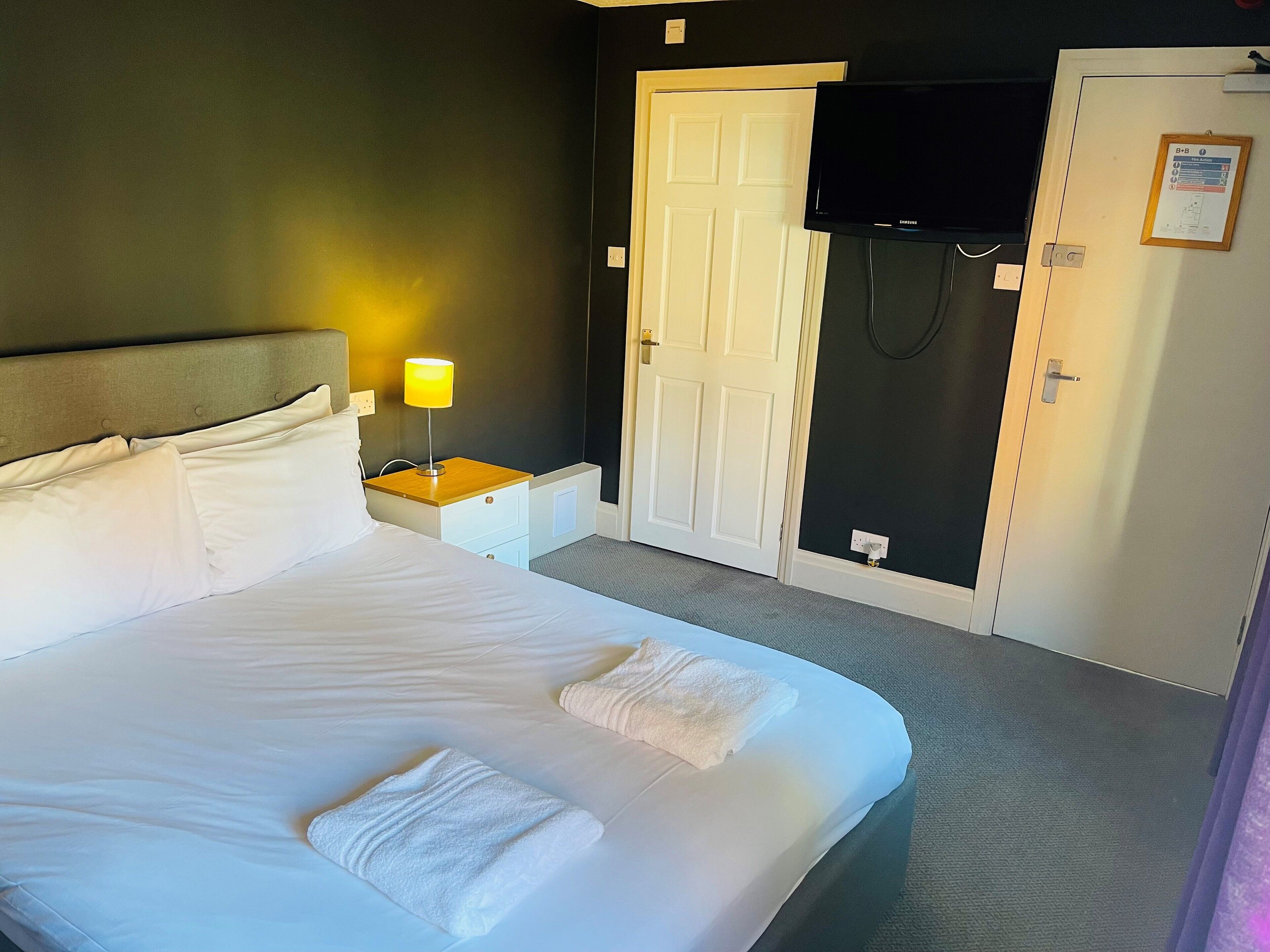 Une chambre d’hôtel avec un lit, une table de chevet avec une lampe, un téléviseur fixé au mur et une porte avec une pancarte.