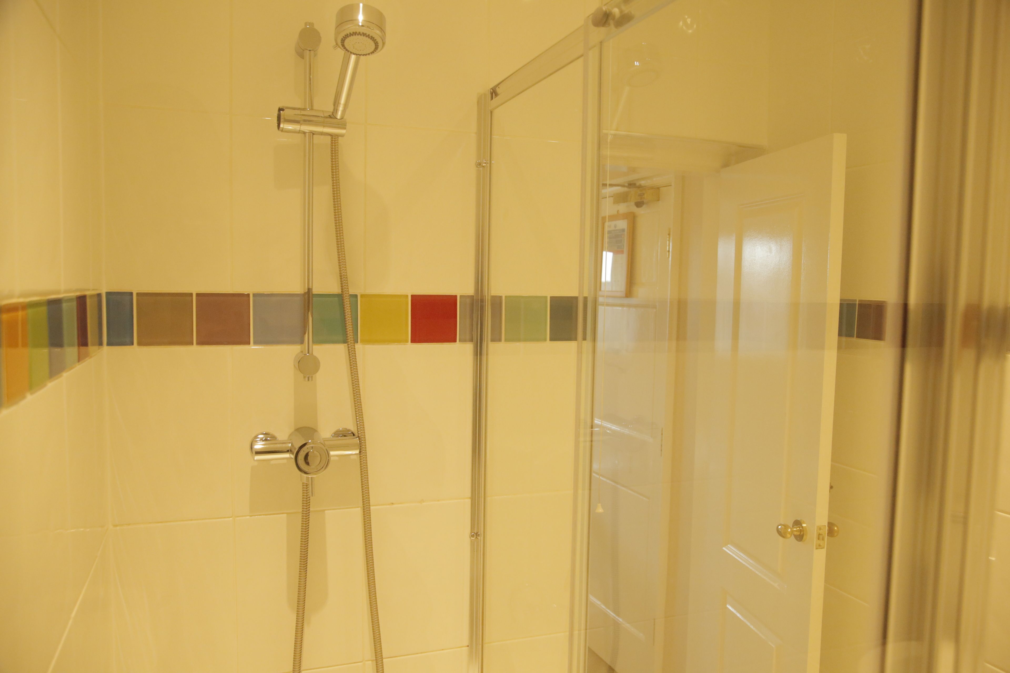A shower with a glass door and a tiled wall with a multicolored border.