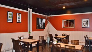 A restaurant interior with wooden tables and chairs, framed artwork on the walls, and a potted plant.