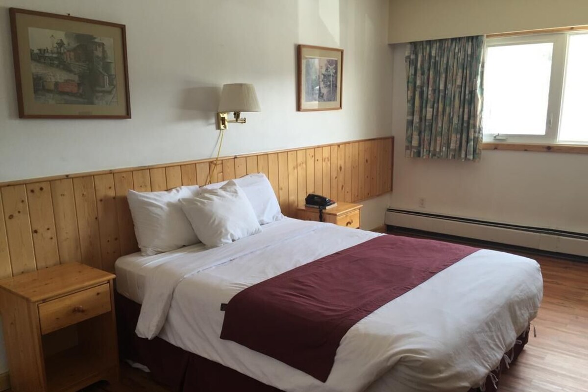 A bedroom with a wooden headboard, a bed with white bedding and a maroon throw, two bedside tables, a nightstand, a lamp, and a window with curtains.