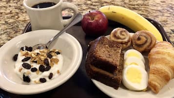 A tray with a bowl of yogurt with raisins, a slice of banana bread, a croissant, a boiled egg, a red apple, and a cup of coffee.