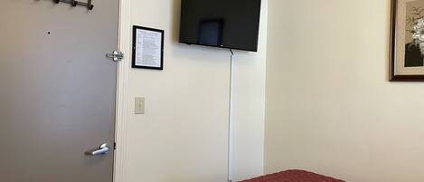 A room with a bed, a door with a coat rack, a mounted television, and a framed picture on the wall.