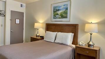 A neatly made bed with a wooden headboard, two bedside lamps, a nightstand with a phone, and a framed landscape painting above the bed.