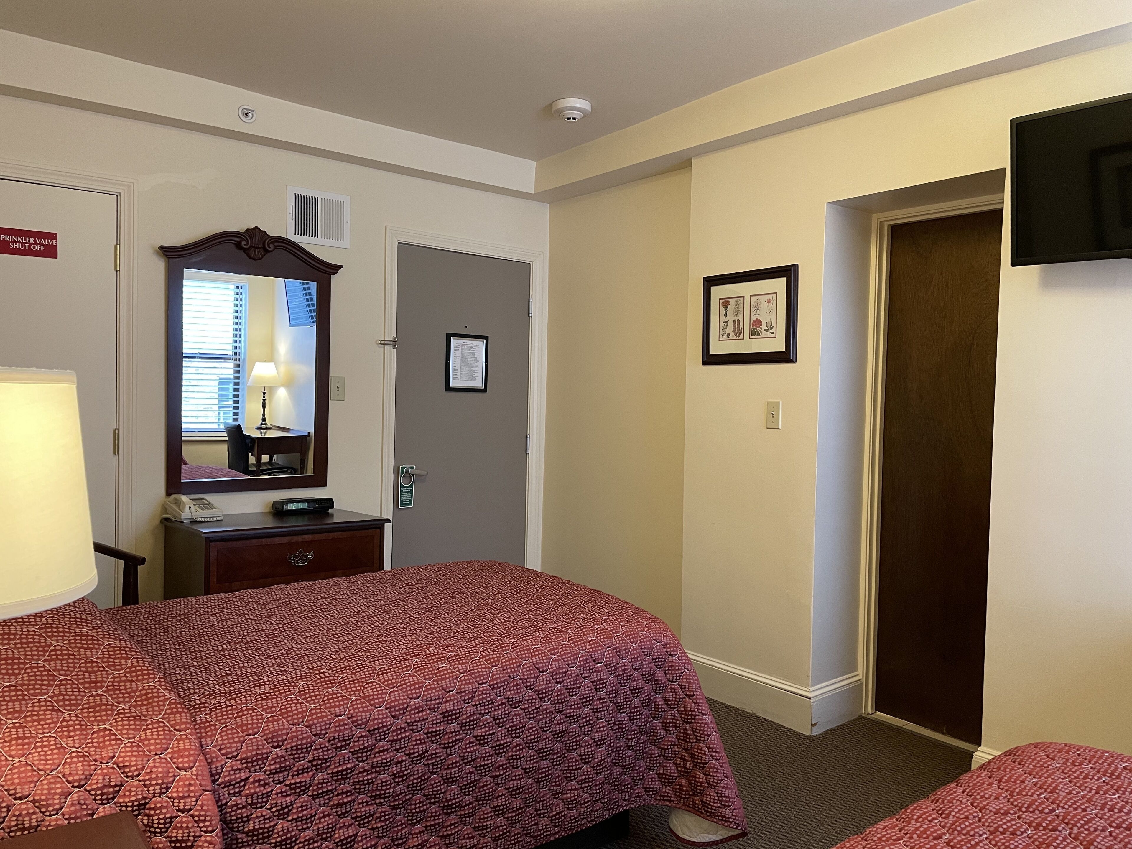 A hotel room with two beds, a desk with a lamp, a mirror, a framed picture on the wall, and a door with a sign.