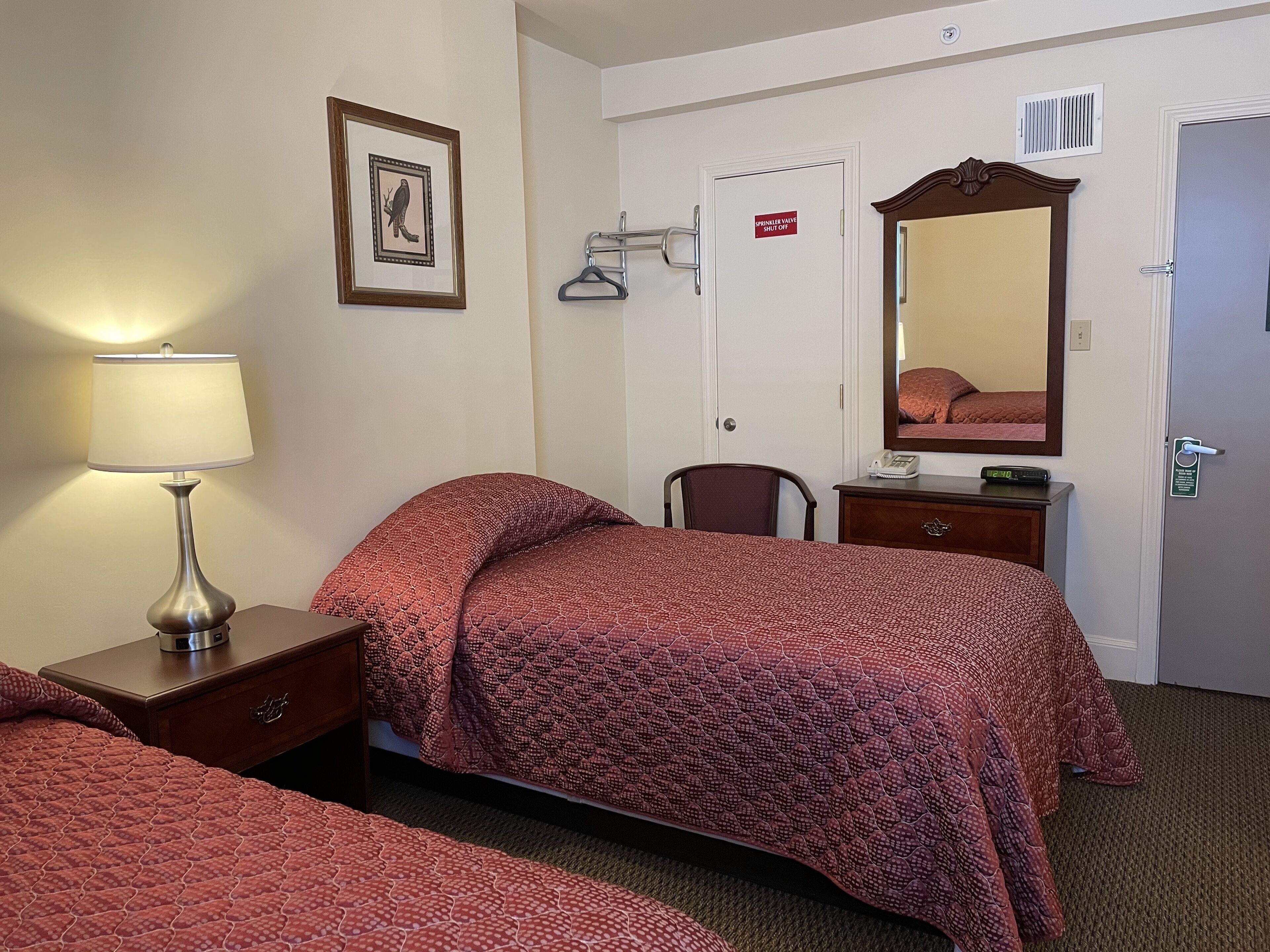 Een hotelkamer met twee bedden, een nachtkastje met lamp, een stoel, een dressoir, een spiegel en een deur met een bordje.