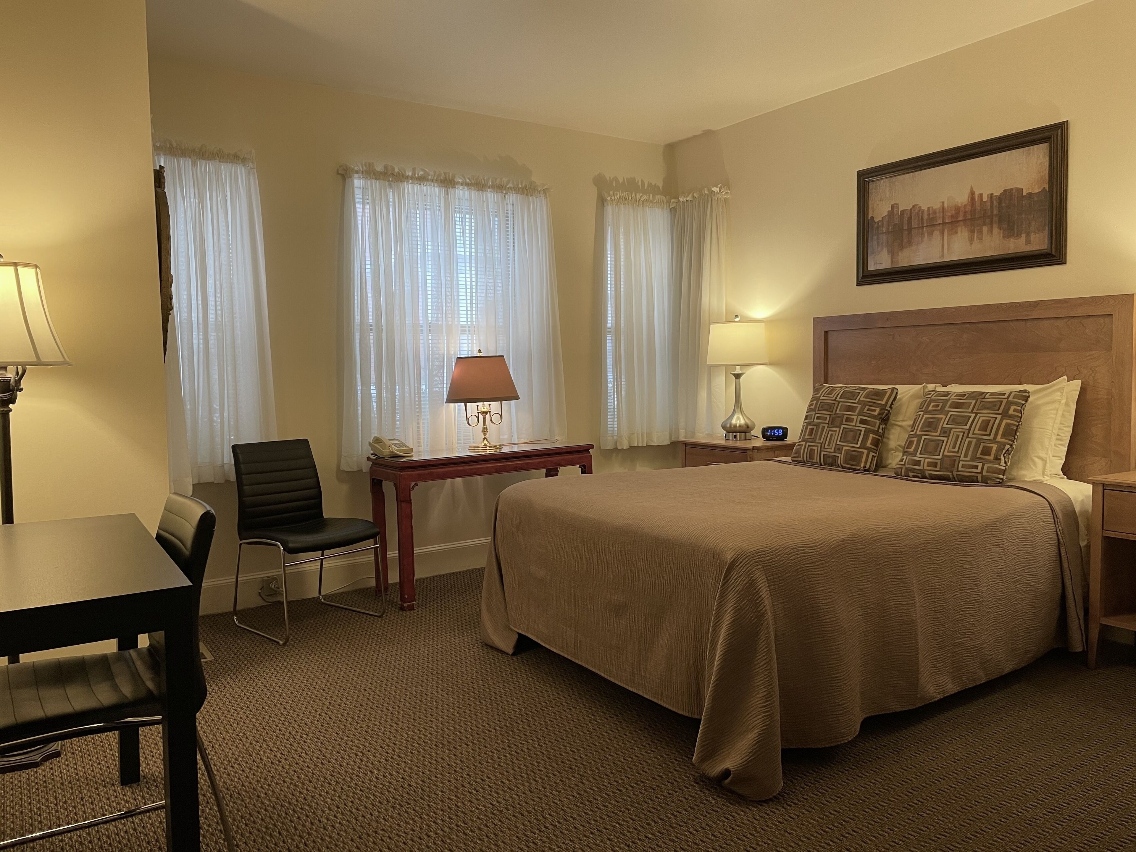 Una habitación de hotel con una cama grande, un escritorio con silla, una mesita de noche con lámpara y un cuadro en la pared.