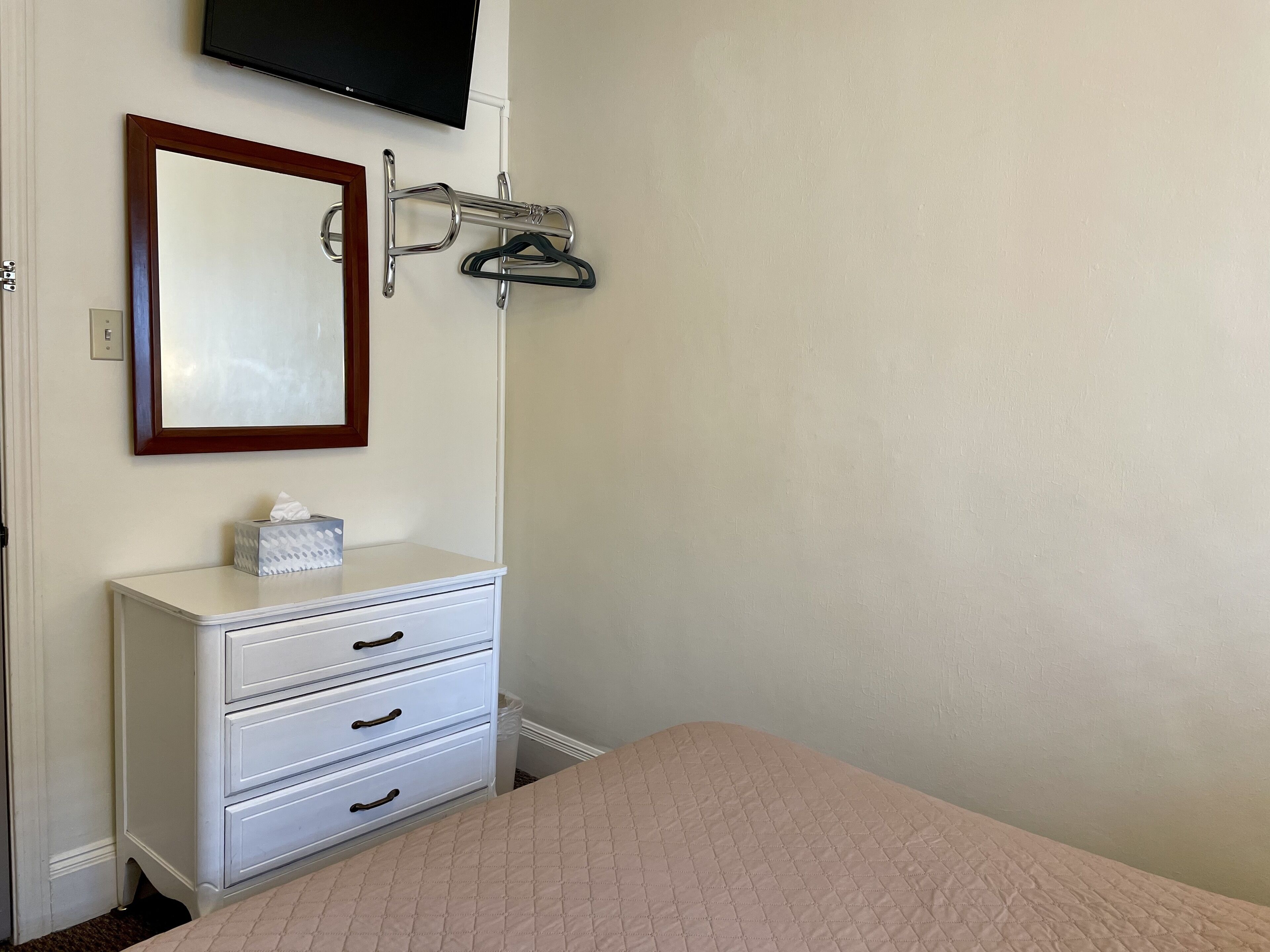 A bedroom with a bed, a dresser with drawers, a mirror, a TV mounted on the wall, and a coat rack with hangers.