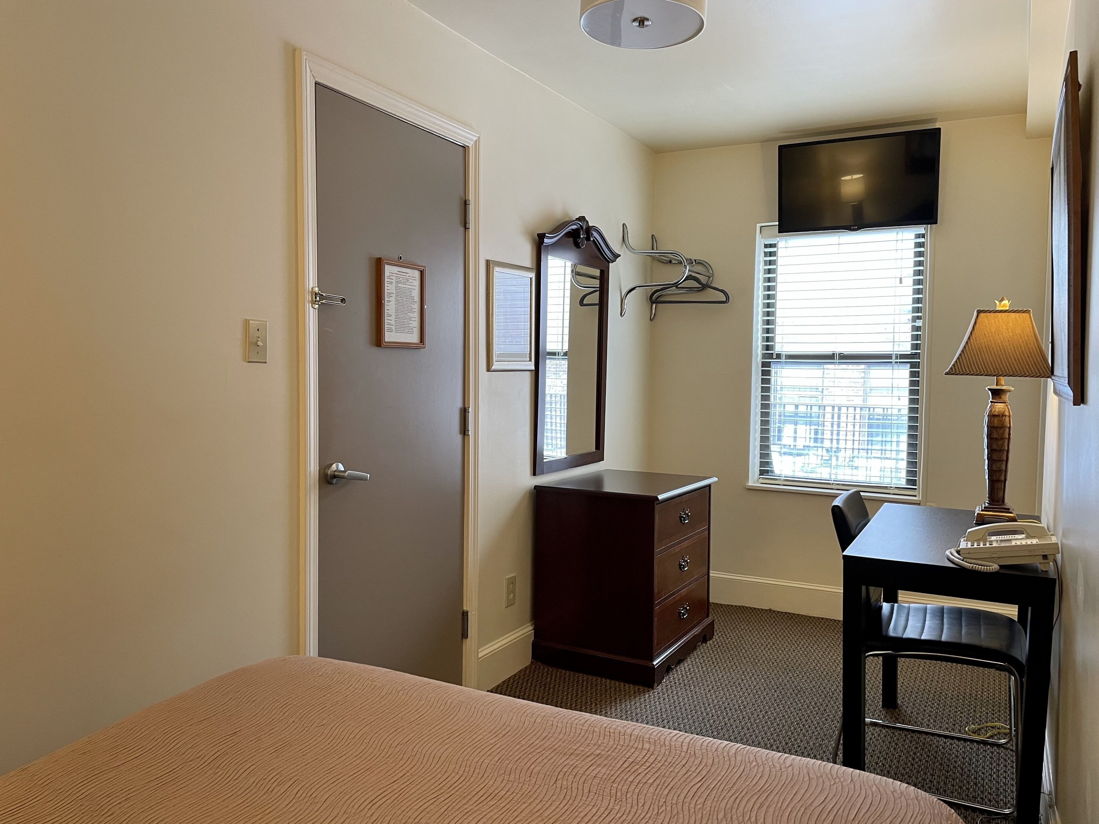A bedroom with a bed, a dresser, a desk with a chair, a mirror, and a window with blinds.