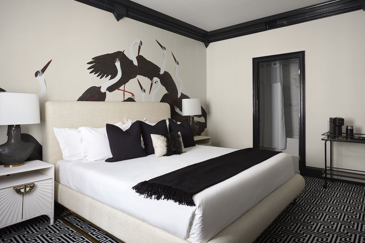A hotel room with a large bed, a nightstand with a lamp, a wall-mounted artwork of birds, and a door with a curtain.