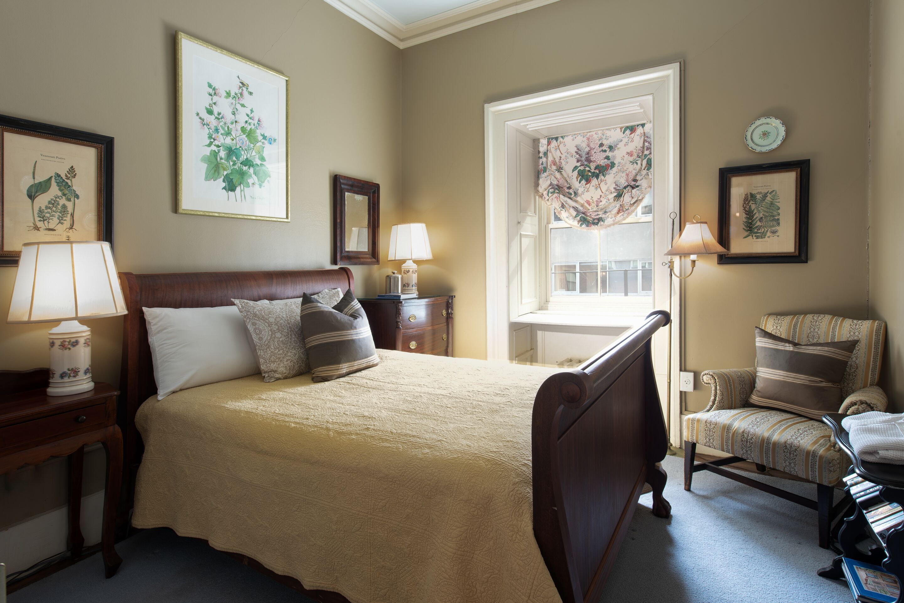 Un dormitorio con una cama grande, una silla, un aparador y una ventana con cortinas florales.