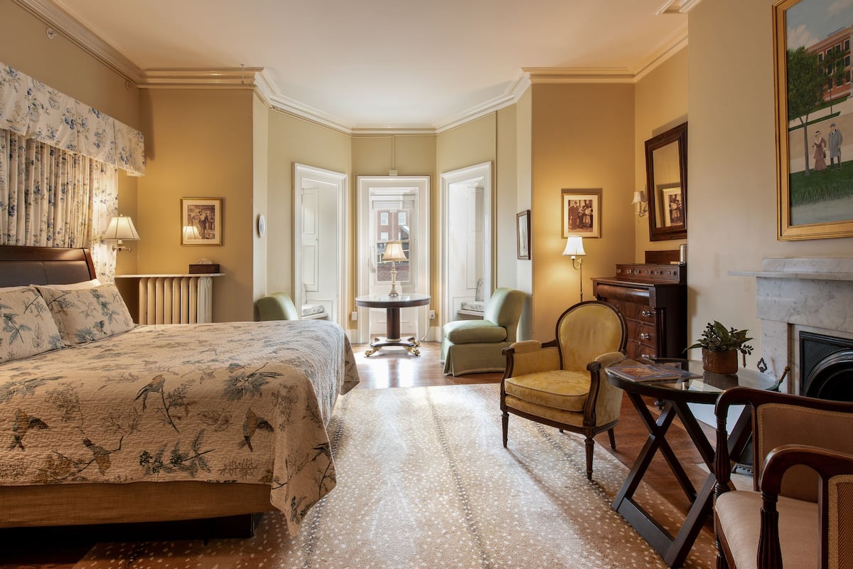 A hotel room with a large bed, a sitting area with a table and chairs, a fireplace, and a view of the street through the windows.