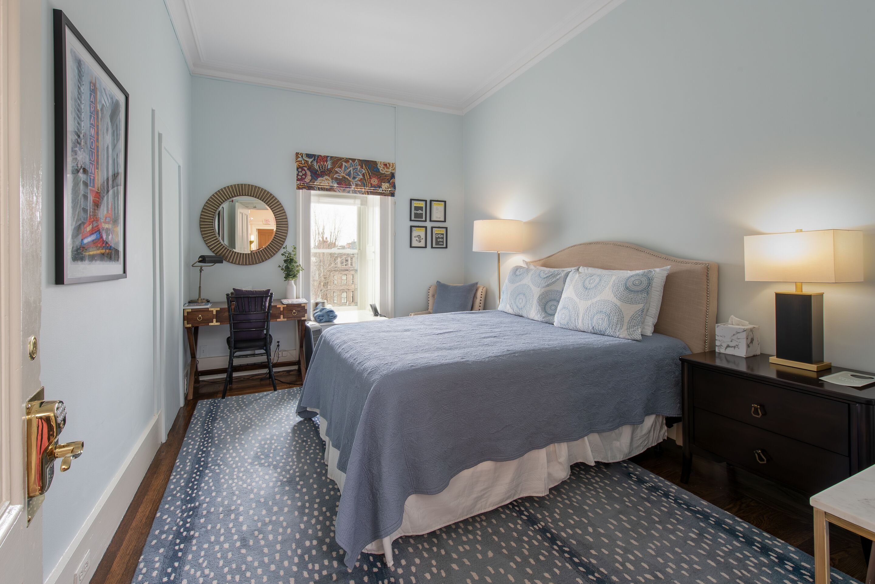 A bedroom with a bed, bedside table, lamp, desk, chair, and a window with a view.