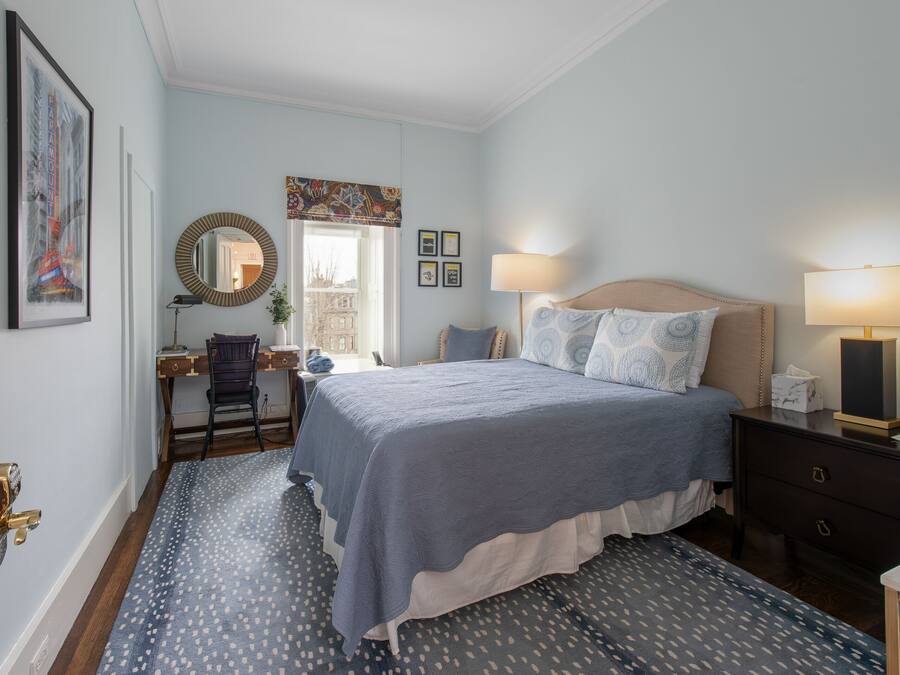 A bedroom with a bed, bedside table, lamp, desk, chair, and a window with a view.