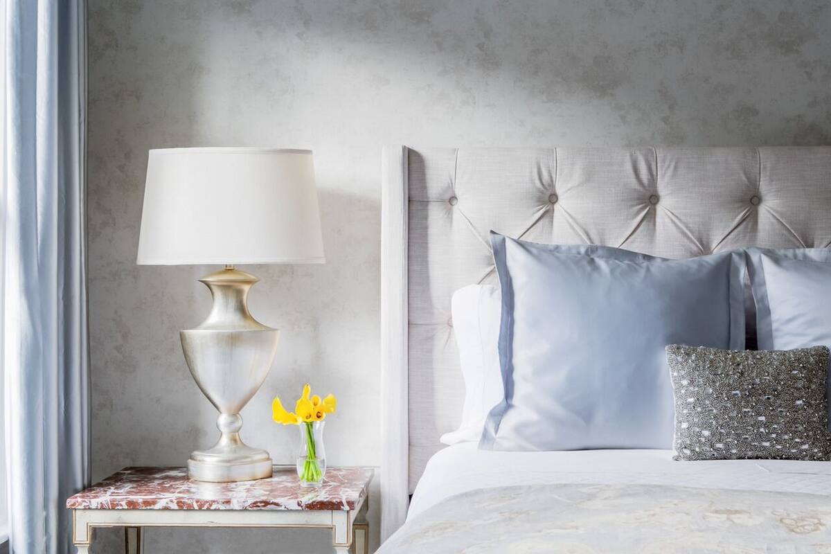 Una cama bien hecha con cabecera capitonada, una mesita de noche con una lámpara y un jarrón con flores amarillas.