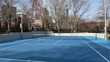 Un court de tennis en plein air, doté d’un filet et entouré d’arbres et d’immeubles.