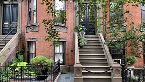 A brick building with a central staircase leading to a black door, surrounded by greenery and a metal fence. - Clarendon Square (Boston)