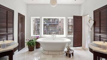 A modern bathroom with a freestanding bathtub, two sinks, and a large window offering a view of the city.