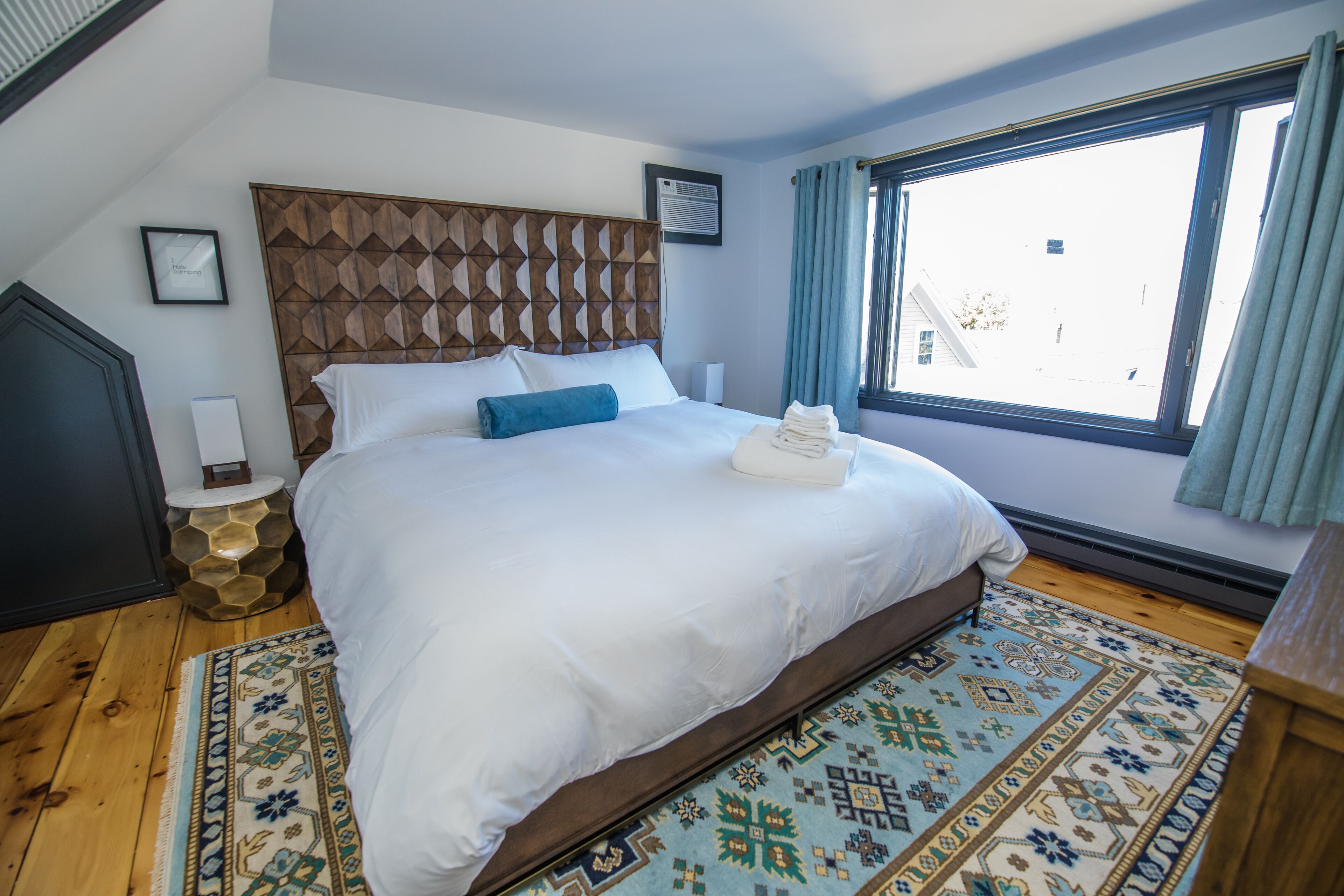 A neatly made bed with a patterned headboard, a wooden floor, and a large window with curtains.