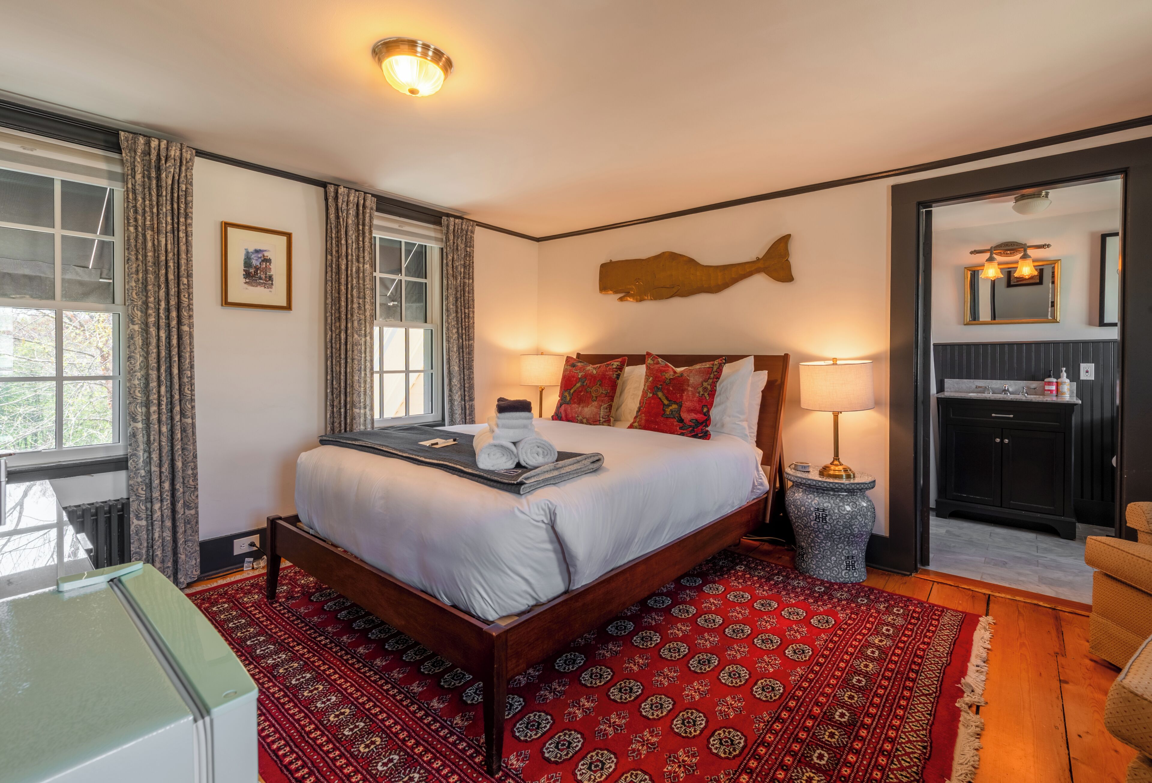 Une chambre d’hôtel avec un grand lit, un plancher en bois, un tapis rouge à motifs, une fenêtre avec des rideaux et une salle de bain visible à travers une porte ouverte.