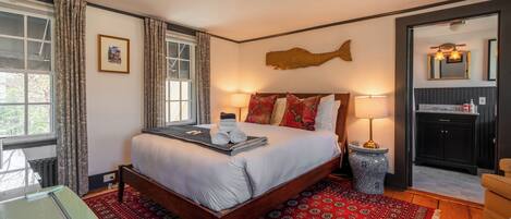 Habitación de hotel con una cama grande, piso de madera, alfombra roja estampada, ventana con cortinas y baño visible a través de una puerta abierta.