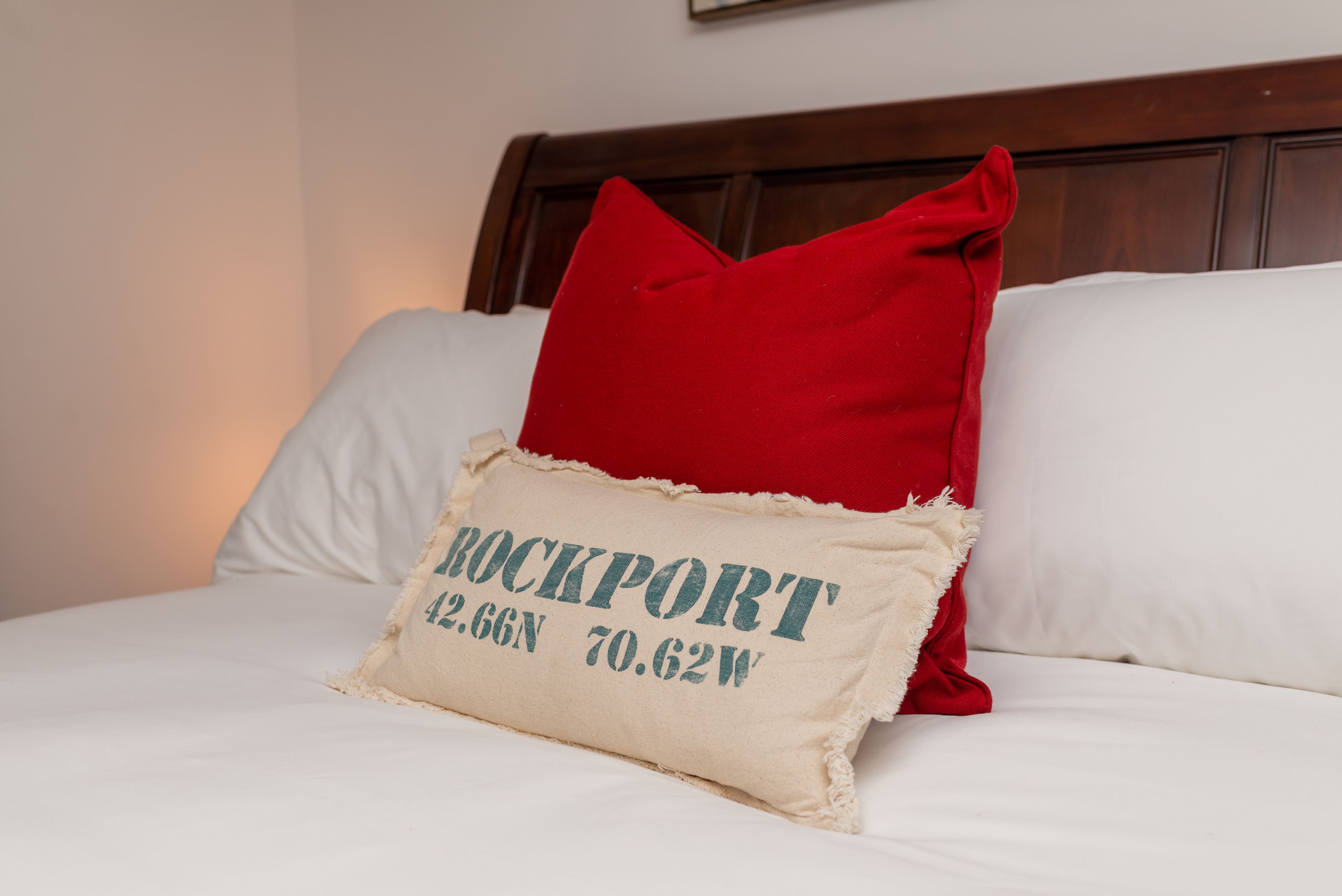 A bed with white bedding, a red pillow, and a beige pillow with coordinates.