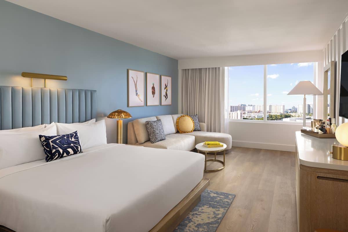 Habitación de hotel con una cama grande, un sofá, una mesita y un televisor. Se ve una ventana con vistas a la ciudad.