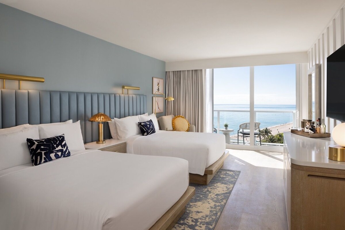A hotel room with two beds, a large window with a view of the beach, a TV, and a small table with a lamp.